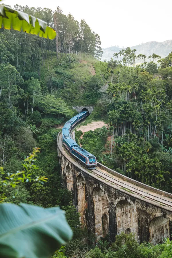 A blue train crossing the Nine Arch Bridge through misty hills near Ella